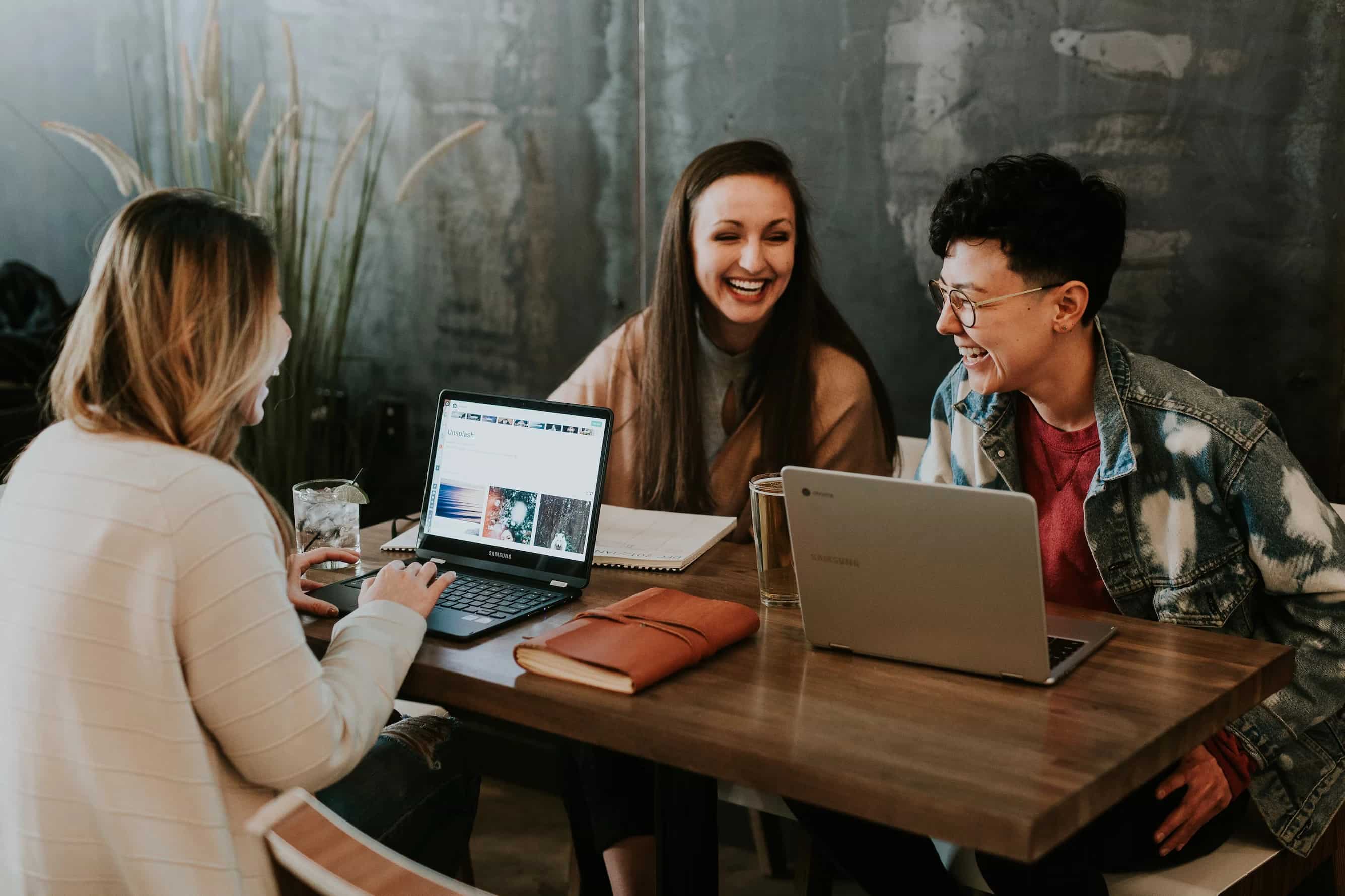 Three people with laptops and notebooks laughing together at a table.